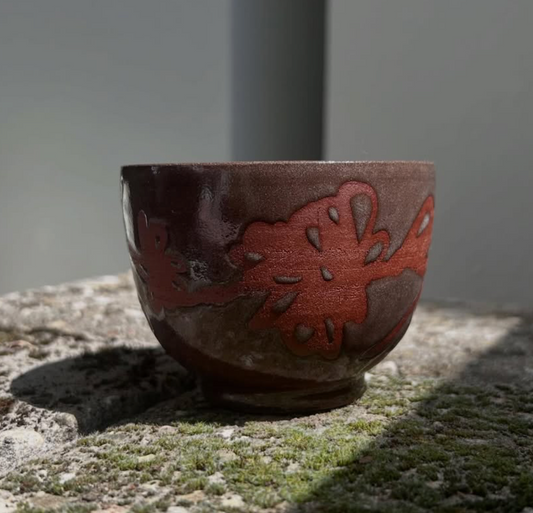 Handgedrehte Keramiknebel Lava-Tasse, ca. 190ml, mit dunkelbrauner, glänzender Glasur und einem erhabenen, leuchtend rotbraunen organischen Muster. Sie steht auf rauem, sonnenbeschienenem Stein und wirft einen definierten Schatten.